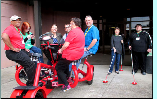 Picture shows people sitting in a circle on a 4-wheeled bicycle, each person has their feet on bicycle pedals.  Beside them are a man and a woman, each holding a cane with a red disk at the bottom, about 10 inches across.